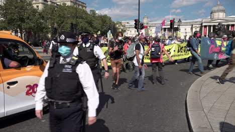 Policías-Con-Máscaras-Faciales-Quirúrgicas-Caminan-Junto-A-Una-Protesta-Por-El-Cambio-Climático-De-La-Rebelión-De-Extinción-Mientras-Sale-De-La-Plaza-Trafalgar-Y-Marcha-Pasando-El-Tráfico-Esperando-En-Un-Semáforo-En-Rojo