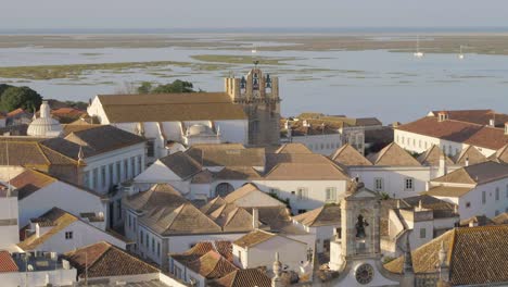 Drone-flies-over-the-old-Traditional-European-village-in-Faro,-Portugal,-revealing-oceanside-rooftops-and-Church-steeple,-Aerial