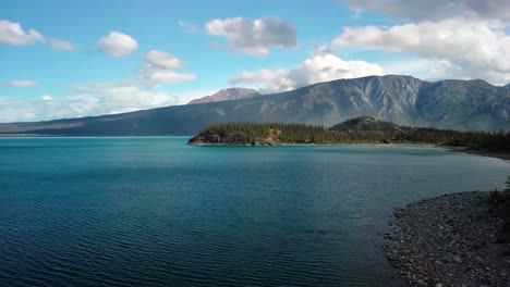 Summertime-seaside-flight-above-Yukon-blue-turquoise-Kluane-lake-shoreline-water-toward-Jacquot-island-and-stunning-mountain-range-on-bright-sunny-sky-day,-Canada,-overhead-aerial-approach