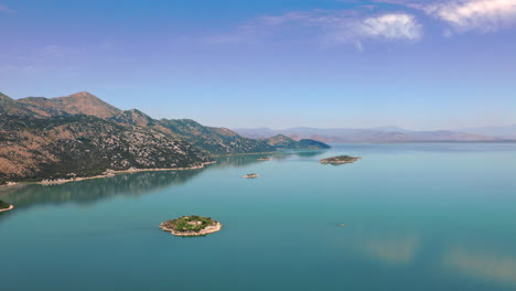 Breathtaking-view-on-lake-Skadar-in-Montenegro-with-its-crystal-clear-blue-water-reflecting-the-sky,-with-tall-mountains-on-the-left-side-and-a-hazy-horizon-in-the-distance,-scenic-aerial-shot-4K