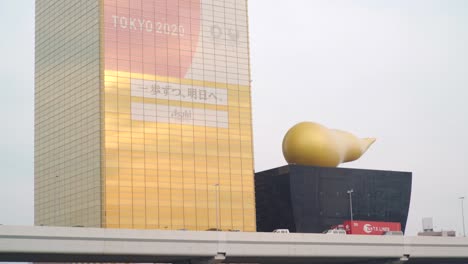 The-Asahi-Breweries-Headquarters-With-Tokyo-Olympic-Digital-Logo-On-The-Building-Exterior-And-The-Giant-Golden-Poop-Sculpture-On-The-Asahi-Beer-Hall-By-The-Sumida-River-In-Asakusa,-Tokyo---wide-shot