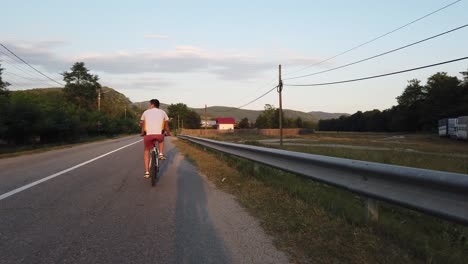 Ein-Junge-Fährt-An-Einem-Sommertag-Bei-Sonnenuntergang-Mit-Dem-Fahrrad-Auf-Einer-Landstraße