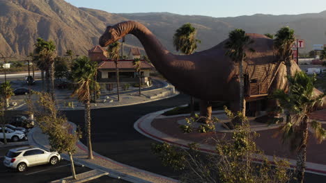 Aerial-view-of-a-Brontosaurus,-one-of-the-Cabazon-Dinosaurs,-World's-Biggest-Dinosaurs,-California,-surrounded-by-palm-trees