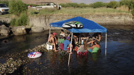 Grupo-De-Jóvenes-Sentados-En-Un-Río-Tranquilo-Celebrando-El-4-De-Julio-Bajo-Una-Pequeña-Carpa-De-Fiesta