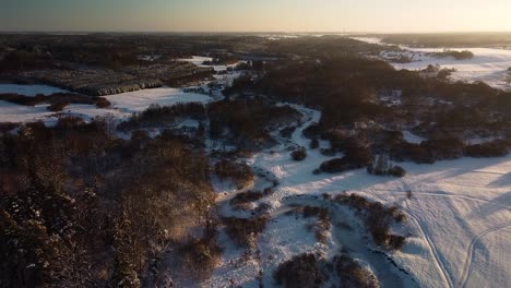 Beautiful-aerial-birdseye-view-of-snow-covered-fields-near-frozen-Sventaja-river-in-sunny-winter-day,-golden-hour,-wide-angle-drone-shot-moving-forward-high-altitude