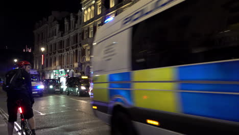 Three-police-vans-with-lights-flashing-drive-at-speed-past-a-cyclist-at-night,-outside-a-theatre-on-Shaftesbury-Avenue-closed-due-to-the-Coronavirus-pandemic