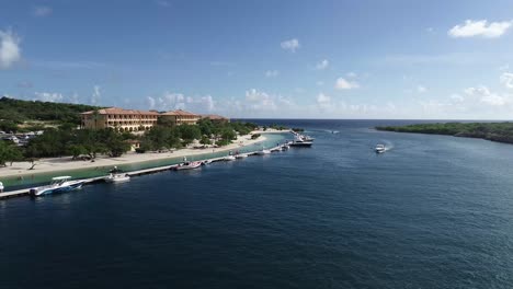 Boat-passing-by-other-boats-moored-by-a-dock-,-beach,-hotel-with-drone-vieuw-with-ocean-view-in-the-background