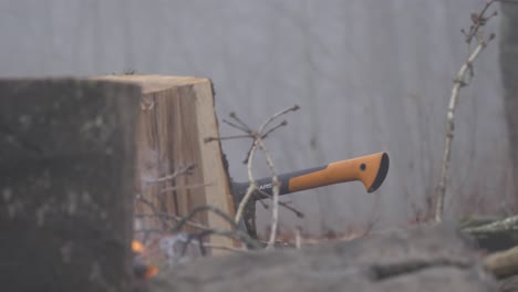 Fiskars-Beil-Neben-Gespaltenem-Holz-Und-Lagerfeuer-Im-Nebligen-Wald-In-Zeitlupe,-Handgehalten