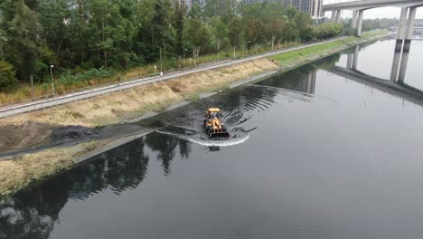 Gelber-Traktor,-Der-Schlamm-Aus-Einem-Kanal-Unter-Einer-Brücke-Schöpft-Und-Reinigt,-Luftbild