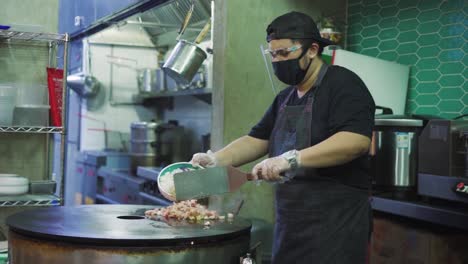 Chef-With-Face-Mask-And-Face-Shield-Pouring-Seafood-And-Rice-On-Hot-Large-Round-Griddle-Pan-Using-Spatula