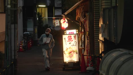 Hombre-Adulto-Con-Mascarilla-En-La-Barbilla-Caminando-En-Un-Callejón-Con-Restaurante-Por-La-Noche-En-Tokio,-Japón