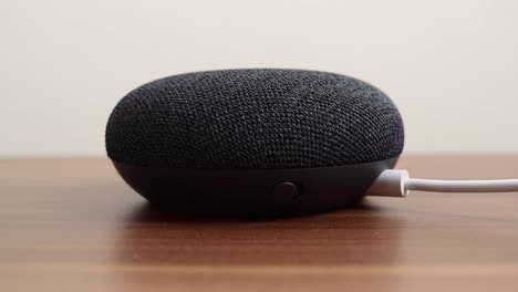 Persona-Presionando-Un-Mini-Altavoz-Inteligente-Google-Nest-Home-Con-Asistente-De-Google-Incorporado-En-Una-Mesa-De-Madera