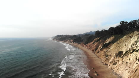 Aerial-drone-shot-over-the-blue-pacific-ocean-waves-off-the-beach-cliffs-on-the-Santa-Barbara,-California-overcast-coastline