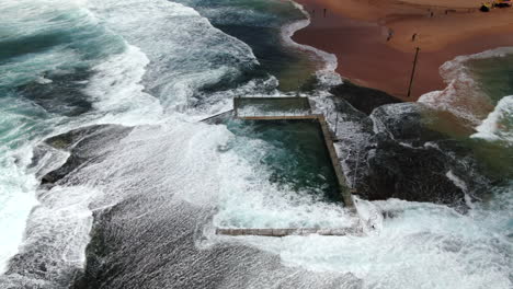 SLOW-MOTION-Aerial-shot-of-waves-and-powerful-ocean-swell-crashing-over-Monavale-Rock-Pool-on-a-beautiful-spring-day