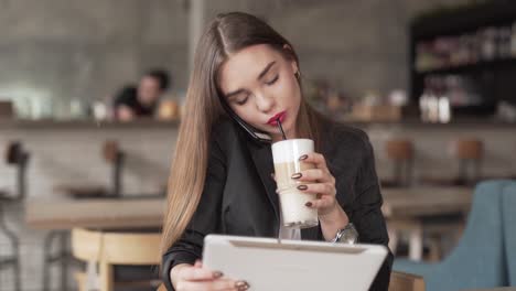 Busy-young-professional-woman-talks-on-her-cellphone,-uses-her-tablet,-and-drinks-coffee-while-sitting-in-a-café