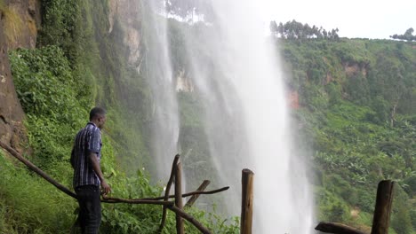 Afrikanischer-Mann-Steht-Hinter-Einem-Tropischen-Wasserfall-Und-Blickt-Ehrfürchtig-Nach-Oben
