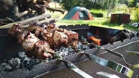 Shashlik-meat-is-bing-grilled-outside-on-the-barbeque,-flames-are-coming,-smokes-rising,-tent-visible-in-the-background---slowmotion,-4K,-30FPS