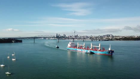Luftaufnahme-Eines-Containerfrachtschiffs,-Das-Von-Schleppern-Unter-Der-Auckland-Harbour-Bridge-Geführt-Wird,-Neuseeland-4k