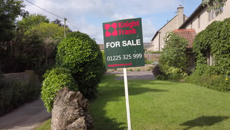 Right-to-Left-Truck-Shot-of-British-‘For-Sale’-Real-Estate-Sign-on-Sunny-Summer’s-Day-with-Rural-Village-Home-in-Background