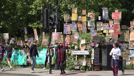 People,-some-in-face-masks,-walk-past-a-huge-memorial-wall-of-cardboard-signs-showing-support-of-healthcare-staff-during-the-Coronavirus-outbreak-line-the-edge-of-a-park-in-the-East-End-of-London