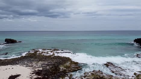 Aerial-drone-flying-from-a-rocky-and-white-sand-beach-towards-a-rough-but-clear-Caribbean-Sea-in-Cozumel,-Mexico