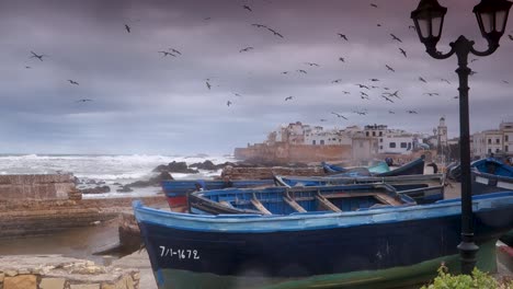 Wide-shot-of-Essaouira,-Morocco-with-the-Atlantic-ocean-slamming-against-the-walls-and-fishing-boats-in-foreground-on-a-stormy-day
