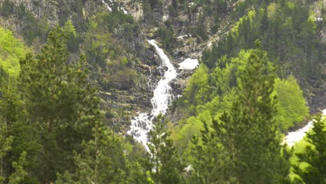 Cascading-waterfalls-on-high-mountainside-seen-through-pine-trees,-slow-motion