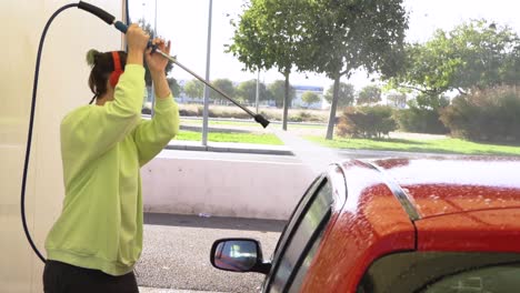 Woman-cleaning-bonnet-red-car-with-pressure-hose-and-listening-to-music-red-headphones