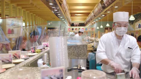 Escena-Dentro-Del-Restaurante-Kaiten-zushi-En-Tokio,-Japón,-Con-El-Chef-Usando-Un-Protector-Facial-Y-Los-Clientes-Separados-Por-Barreras-De-Láminas-De-Plástico-Para-Evitar-La-Propagación-Del-Coronavirus---Tiro-Medio