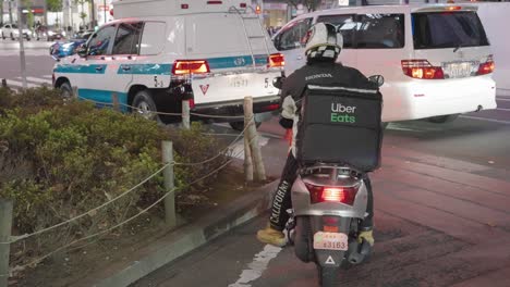 Uber-Come-Motociclista-De-Entrega-De-Alimentos-Esperando-En-El-Cruce-De-Shibuya-En-Tokio,-Japón-Por-La-Noche