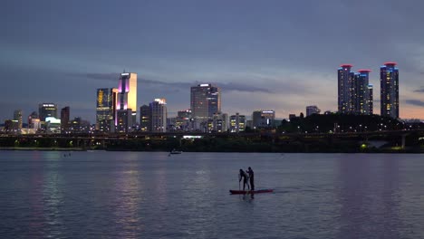 Silhouette-of-Unrecognizable-Man-and-Woman-Paddling-on-SUP-on-Han-River-at-Twilight,-Olympic-daero-Highway,-Trade-Tower,-Asem-Tower-on-Purple-Sky-at-Sunset-Background,-Static-4k