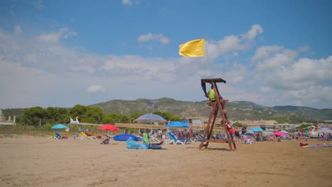 Socorrista-De-Playa-En-Su-Cabaña-Mirando-A-Los-Nadadores-Con-Una-Bandera-Amarilla-Ondeando-A-Cámara-Lenta