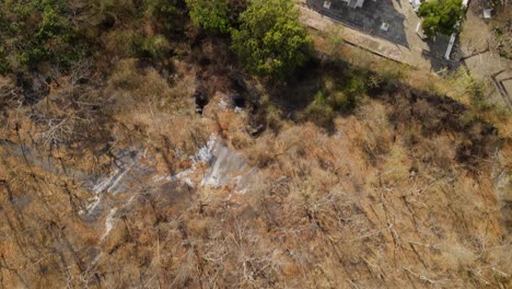 Wat-Phra-Phutthabat,-Saraburi,-Thailand,-4k-aerial-footage-from-above-the-dry-and-brown-forest-during-summer-towards-the-temple-complex,-road-on-the-right,-green-trees-at-the-garden