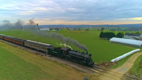 An-Aerial-View-of-an-Antique-Restored-Steam-Train-and-Passenger-Coaches-Approaching-With-Smoke-and-Steam-Along-Green-Farmlands-and-Fields-as-Seen-by-a-Drone