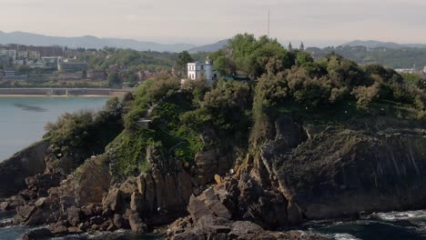 Lonely-white-building-on-rocky-island-with-flying-birds-and-San-Sebastian-in-background,-cinematic-aerial-view