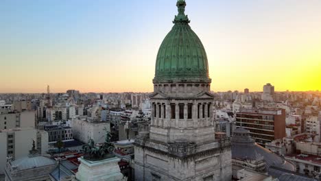 Cinematic-aerial-pan-shot-around-bronze-plated-dome-shaped-cupola-at-the-historical-building,-Palace-of-the-Argentine-National-Congress-surrounded-by-populated-cityscape-and-sun-setting-at-horizon