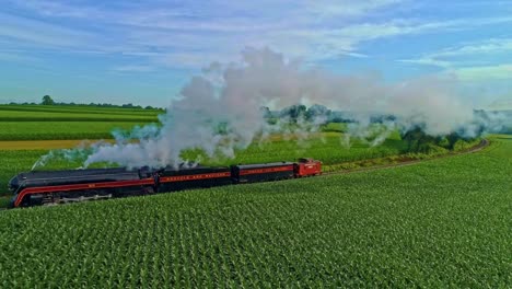 Paisaje-Aéreo-De-Tierras-De-Cultivo-Y-Un-Motor-De-Vapor-Antiguo-Pasa-A-Través-De-Los-Campos-De-Maíz-Y-Corre-Paralelo-En-Una-Mañana-De-Verano-Temprano