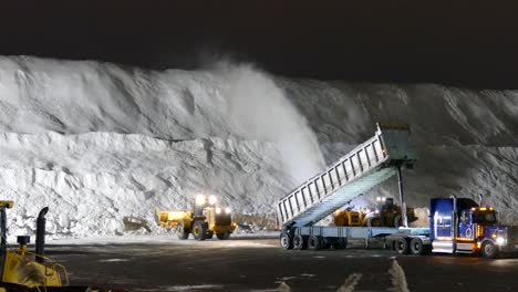 The-truck-is-shoveling-snow-from-the-trailer,-the-yellow-excavators-are-clearing-the-snow,-blowing-snow-in-a-big-pile