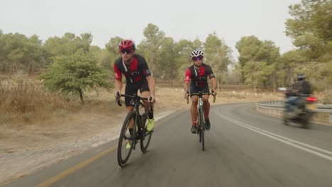 Dos-Ciclistas-De-Carretera-Profesionales-Cabalgando-Por-La-Carretera-Vacía-Del-Bosque