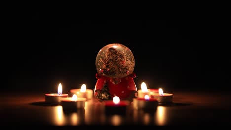 Isolated-Christmas-snow-globe-sphere-with-a-miniature-santa-in-a-winter-snow-fall-inside,-surrounded-by-candles-over-a-black-background