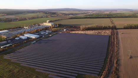 Drone-shot-of-solar-panels-producing-green-electricity,-surrounded-by-agricultural-fields-on-three-sides-and-a-factory-on-one-side-on-a-bright-sunny-day