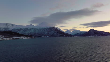 Sunset-brings-gold-sky-behind-snowy-mountains-in-Arctic,-ocean-aerial