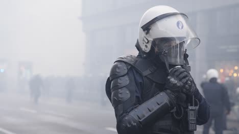 Ein-Einzelner-Polizist-In-Voller-Kampfmontur-Setzt-Während-Eines-Rauchigen-Aufstands-Eine-Gasmaske-Auf-Sein-Gesicht,-Zeitlupe