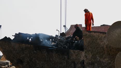 Two-Workers-On-Dismantled-Ship-Hull-At-Gadani-With-Smoke-From-Acetylene-Torch-Going-Past