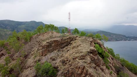 Drone-Aéreo-Que-Se-Eleva-Sobre-Una-Torre-De-Telecomunicaciones-Roja-Y-Blanca-En-La-Cima-De-Una-Montaña-Rocosa-En-Las-Afueras-De-La-Capital-Dili,-Timor-Leste