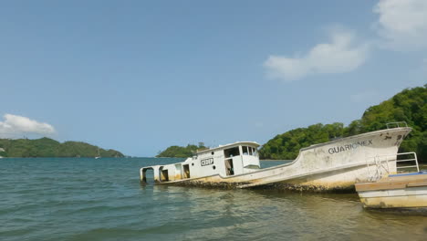 Rocas-Cubiertas-De-Musgo-En-La-Orilla-De-La-Playa-Con-Un-Barco-Naufragado-En-La-Distancia-En-Samana,-República-Dominicana