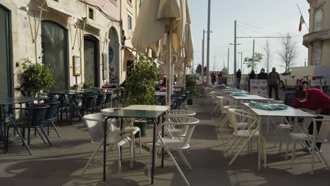 Vista-De-Una-Mesa-Y-Sillas-Vacías-Fuera-Del-Restaurante-En-Lisboa-Con-El-Viento-Soplando-Sobre-Sombrillas