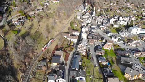 Antena:-Tren-Rojo-Y-Blanco-En-Los-Alpes,-Recorriendo-La-Ladera-De-Una-Montaña-Y-Saliendo-De-Un-Pueblo-Alpino