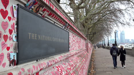 People-walk-past-the-plaque-and-photos-and-the-thousands-of-red-painted-hearts-with-messages-to-lost-loved-ones-during-the-pandemic-at-The-National-Covid-Memorial-Wall
