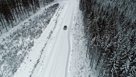 Aerial-tracking-shot-of-car-driving-on-snowy-road-in-forest-landscape-during-winter-day